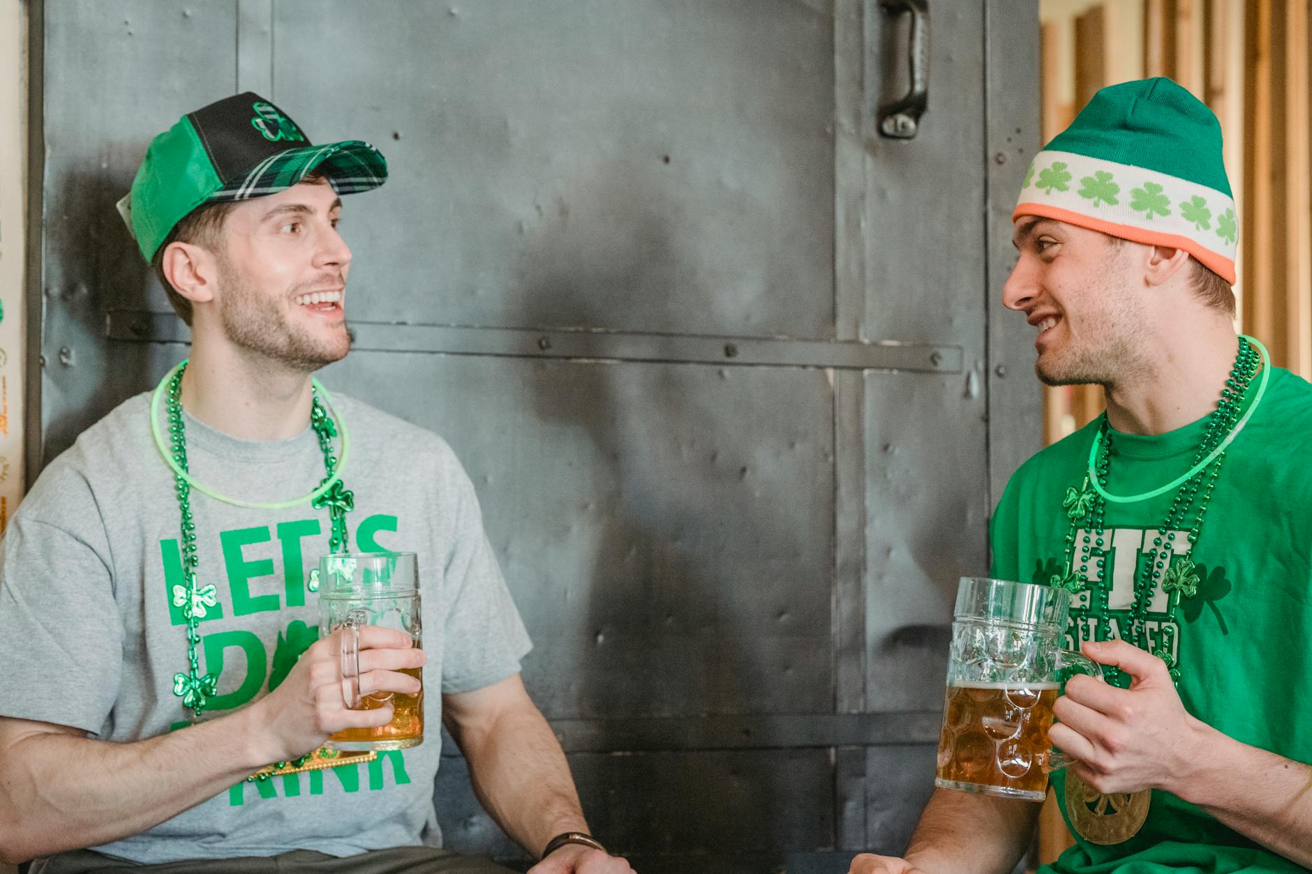 Cheerful unshaven friends in green hat with symbols of Saint Patricks Day wearing neon green glow stick and necklace decorated with shamrock and green beads talking and drinking ale