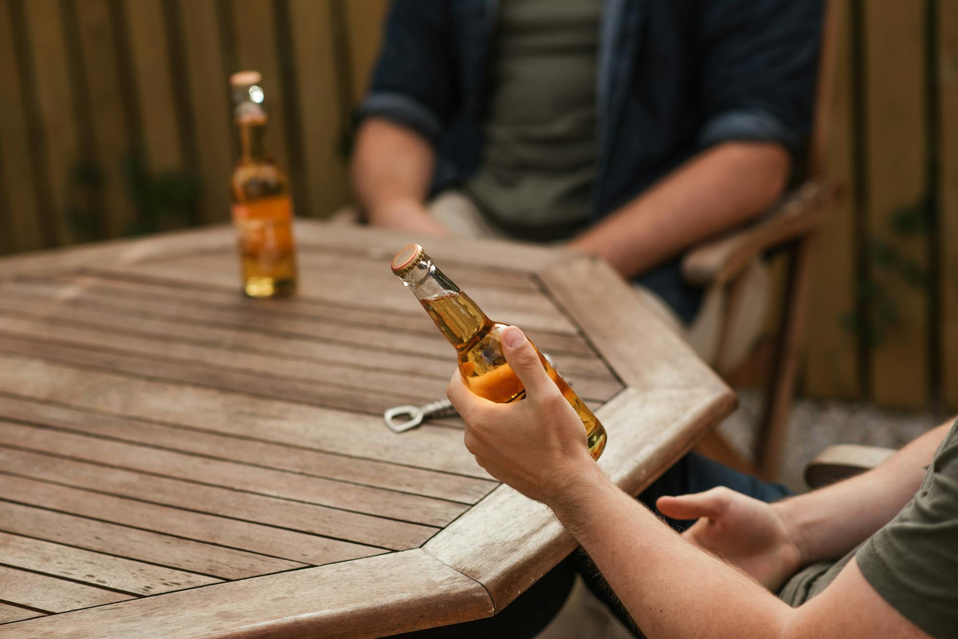 Crop anonymous male partners with glass bottles of alcoholic drink sitting at wooden table on weekend