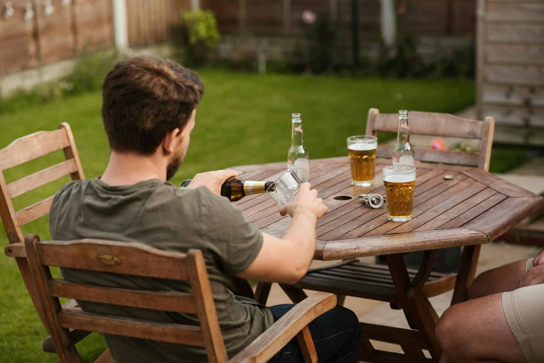 Back view of anonymous male filling glass mug with beer from bottle while chilling with friend at backyard