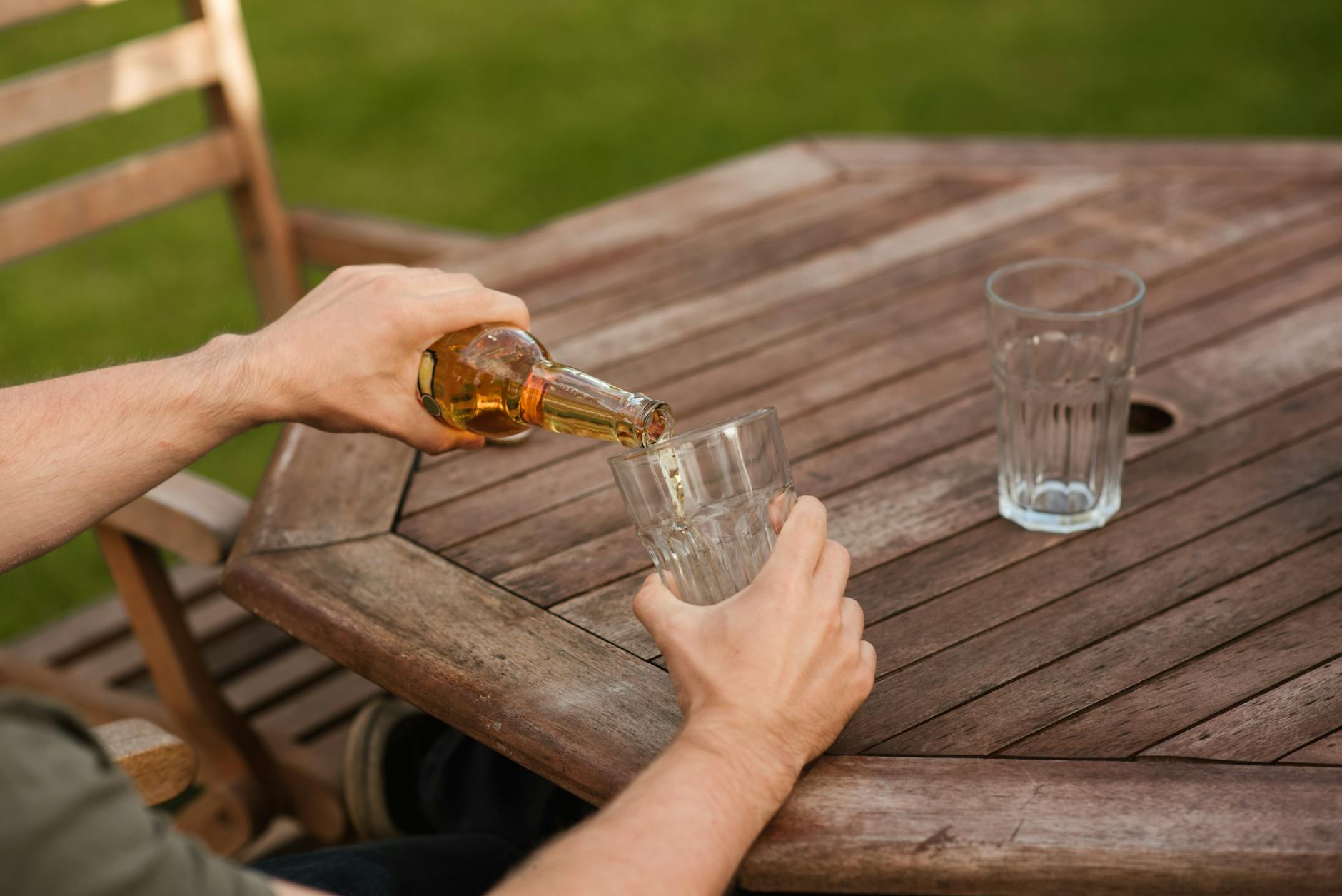 From above crop anonymous male sitting at wooden table and pouring cold beer into glass cup at weekend in yard