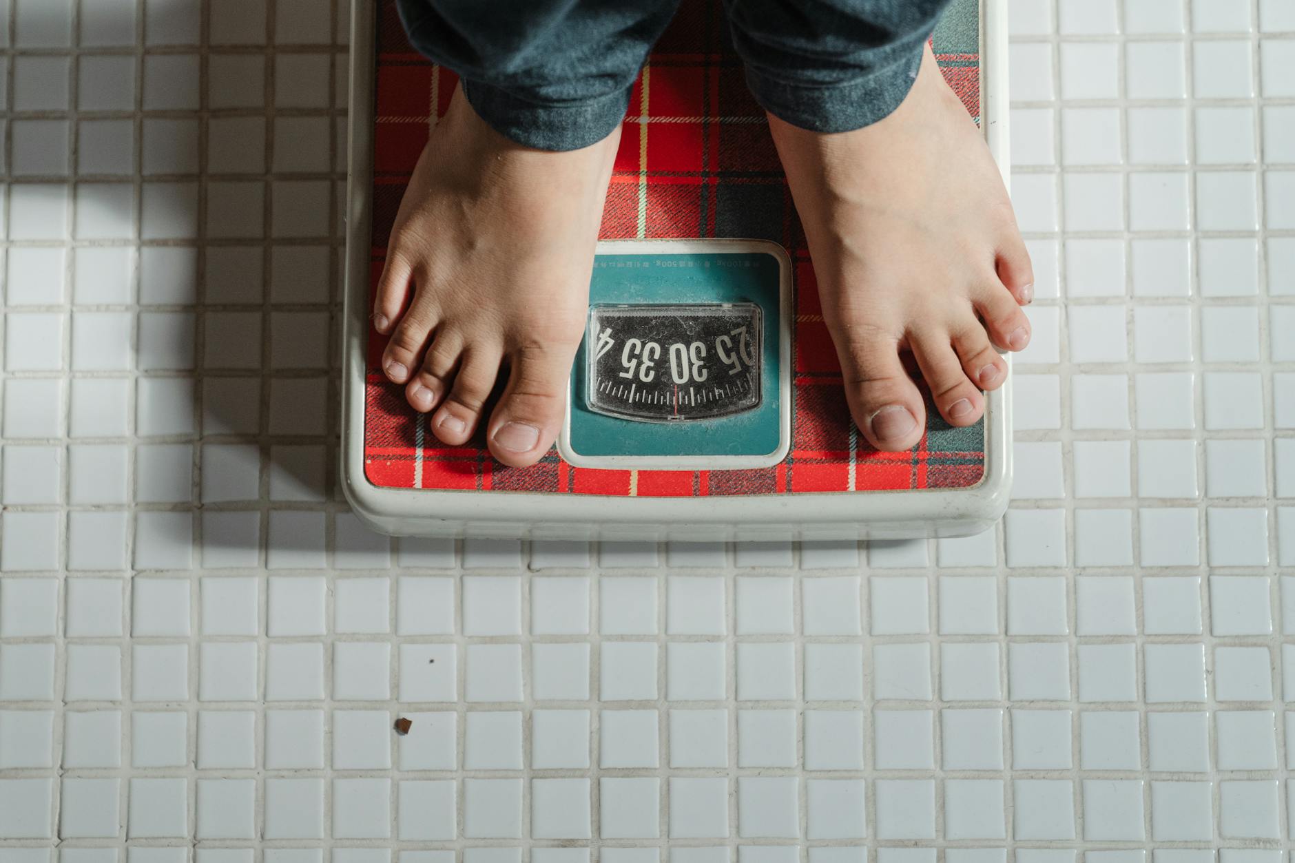 From above crop anonymous barefoot child in jeans standing on weigh scales on tiled floor of bathroom