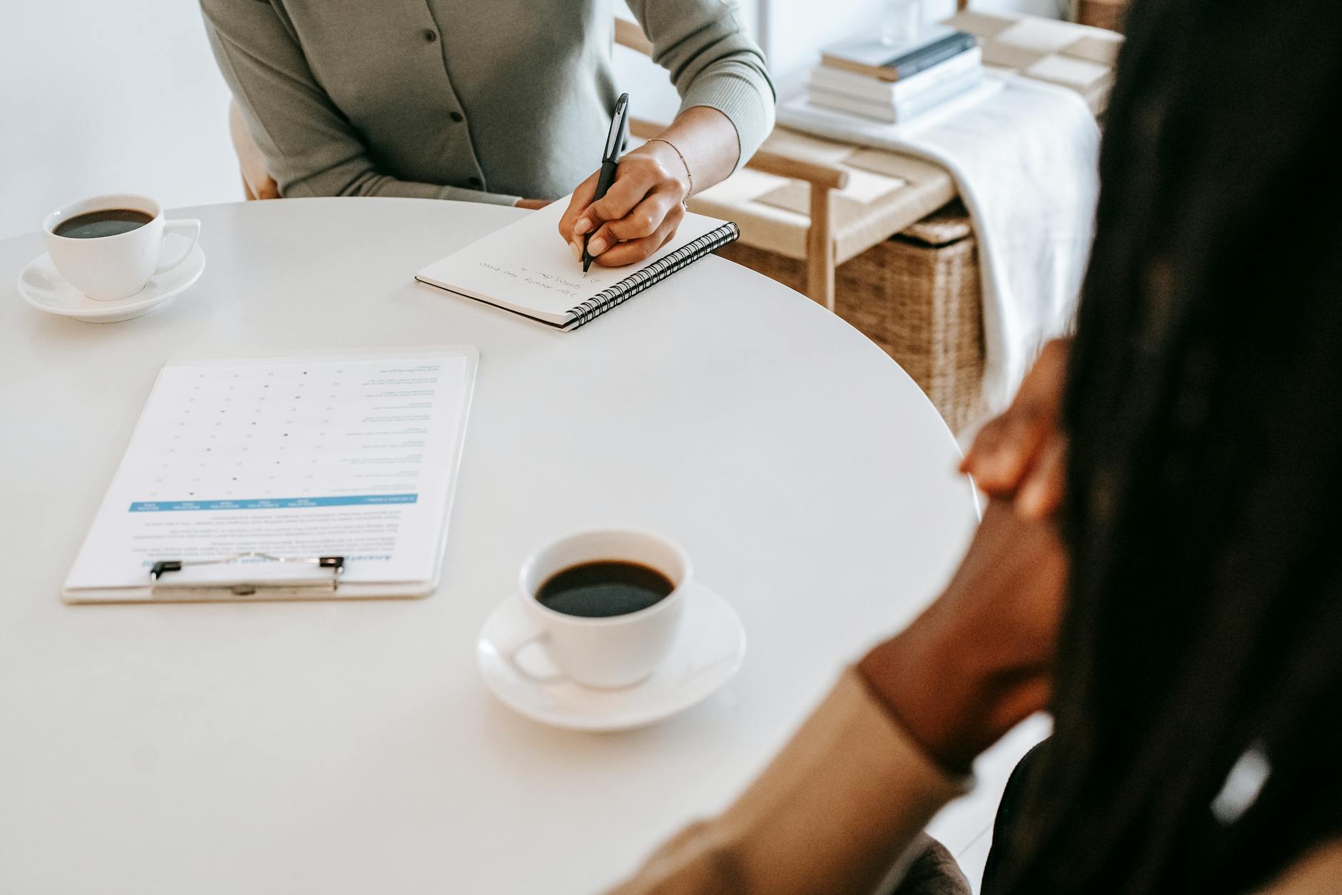 From above crop multiracial female interviewer or psychologist in formal wear asking questions and taking notes in planner while talking to black man and sitting together at table with coffee