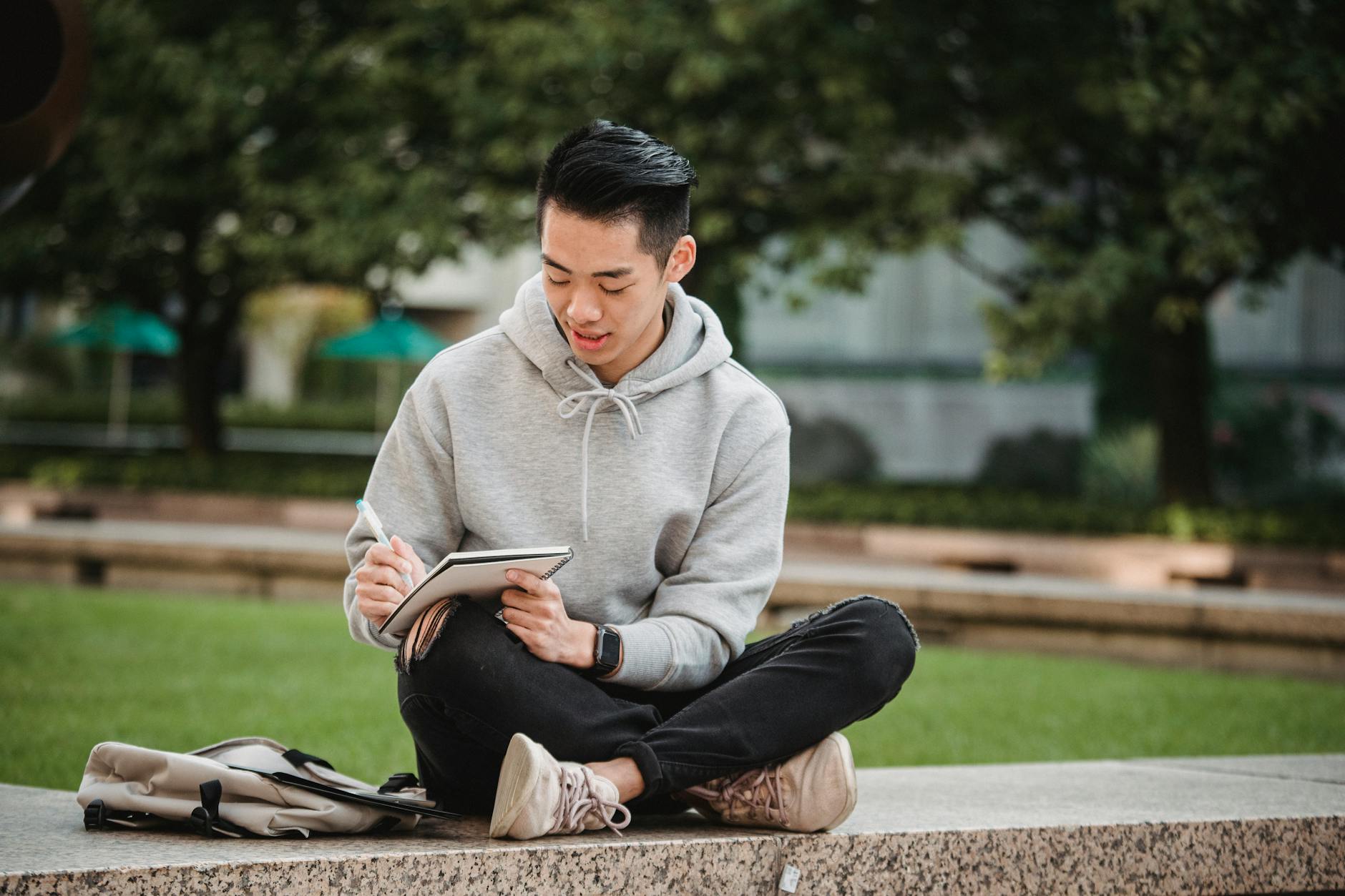 Full body of concentrated Asian male in gray hoodie and ripped black jeans sitting on stone border with legs crossed and writing in notepad